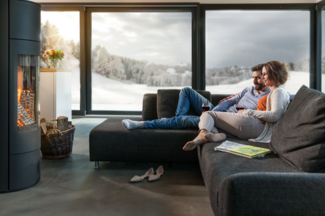 Paar entspannt auf einem modernen Sofa vor einem knisternden Kaminfeuer, mit Blick durch grosse Fenster auf eine verschneite Landschaft.