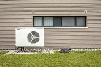 Aussenansicht eines modernen Hauses mit Holzfassade; vor der Wand steht eine Luft‑Wasser‑Wärmepumpe, daneben ein autonomer Mähroboter auf der Rasenfläche.